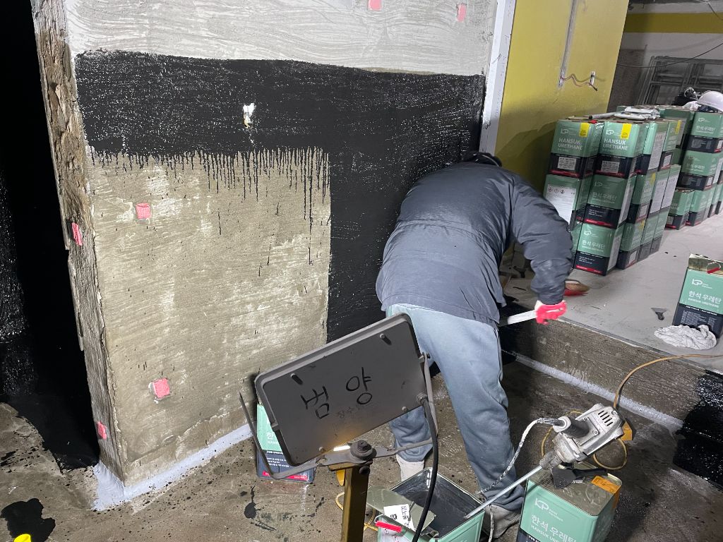 우레탄 중도 도포 — 작업자가 벽면에 우레탄을 도포 중. 뒤쪽에 한석 우레탄 자재통이 쌓여있다