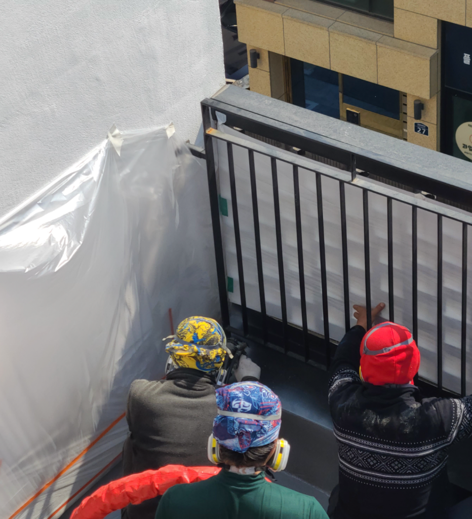 벽면을 비닐로 양생한 뒤 작업자 3명이 난간 앞에서 스프레이 작업 중