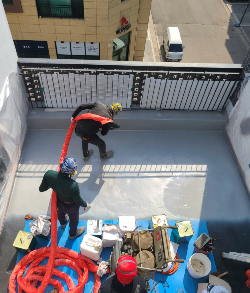 위에서 내려다본 폴리우레아 시공 중 전경 — 빨간 호스와 장비, 작업자, 회색 도포면이 한눈에 보인다