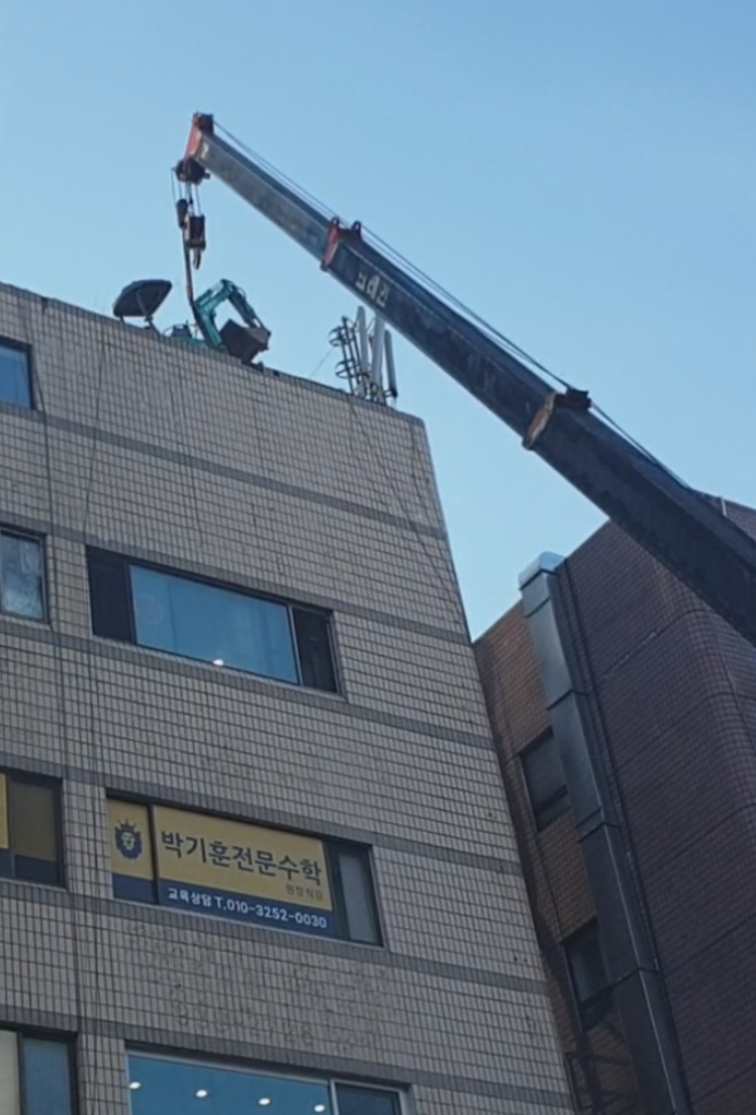 크레인이 미니 굴삭기를 건물 옥상으로 올리는 장면 — 1층 박기훈전문수학 간판이 보이는 현장
