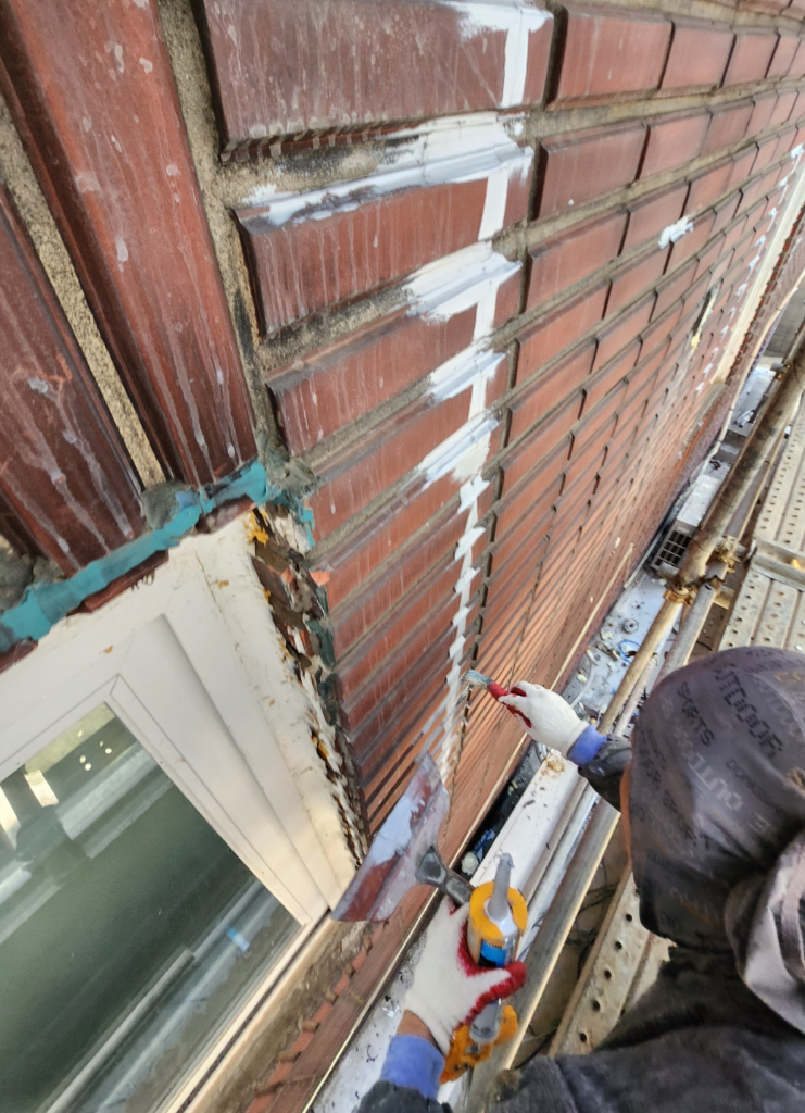 작업자가 코킹건으로 벽돌 줄눈에 실란트를 충전하는 모습 — 수직·수평 줄눈 모두 코킹 진행 중