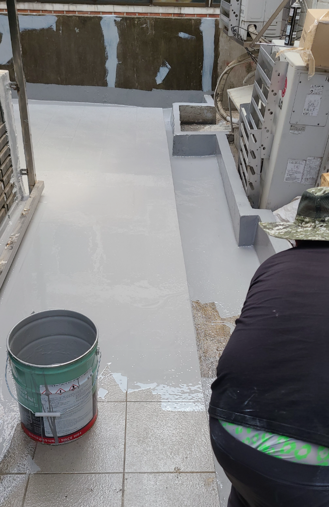 바닥 우레탄 중도 시공 중 — 작업자가 녹색 페인트통의 방수재를 타일 위에 도포하고, 벽면 하단부까지 방수재가 올라간 상태