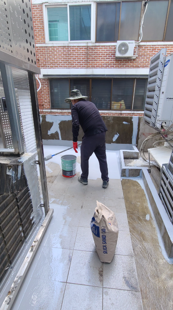 작업자가 바닥에 우레탄 방수재를 도포하는 모습 — 녹색 페인트통과 규사(SILICA SAND) 포대가 놓여 있고, 타일 위에 흰색 방수재를 바르는 중