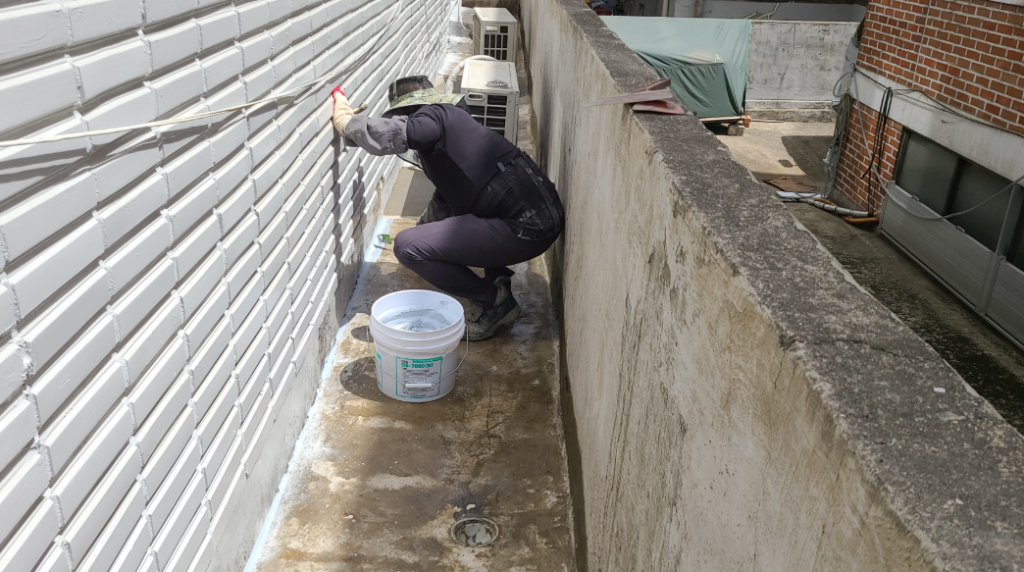 계단실 통로에서 작업자가 벽면에 방수재를 도포하는 모습 — 흰색 페인트통을 옆에 두고, 좁은 통로 벽면에 롤러로 방수 코팅 작업 중